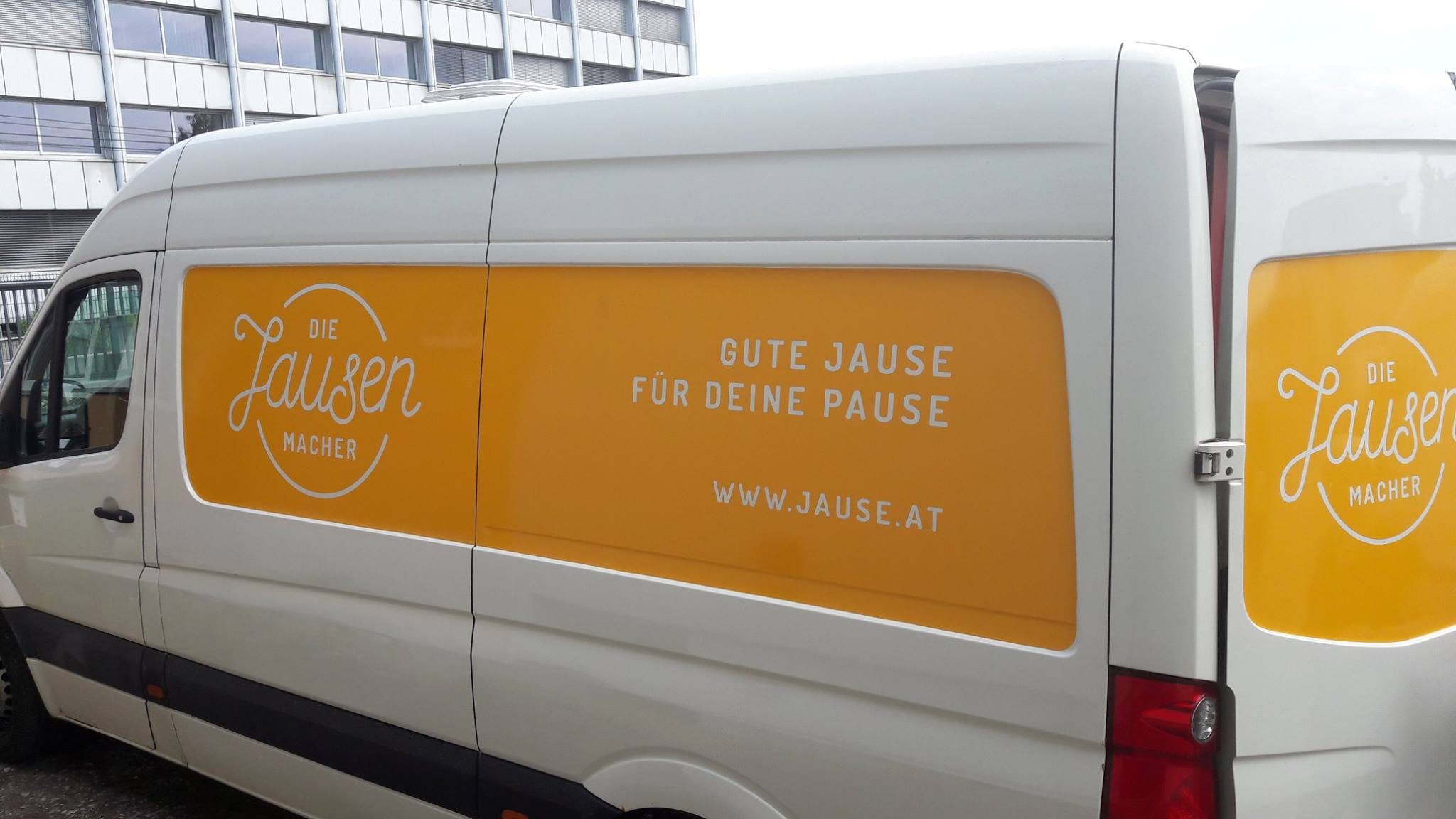 Weißer Lieferwagen der Jausenmacher mit orangem Seitenfeld und der Aufschrift „Gute Jause für deine Pause“